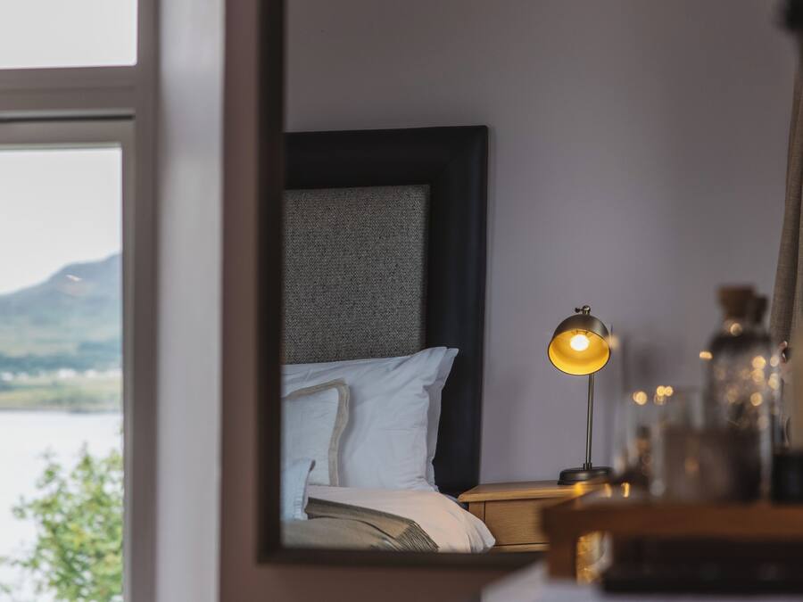 Un dormitorio con cama, mesita de noche, lámpara y vistas a un cuerpo de agua y colinas.