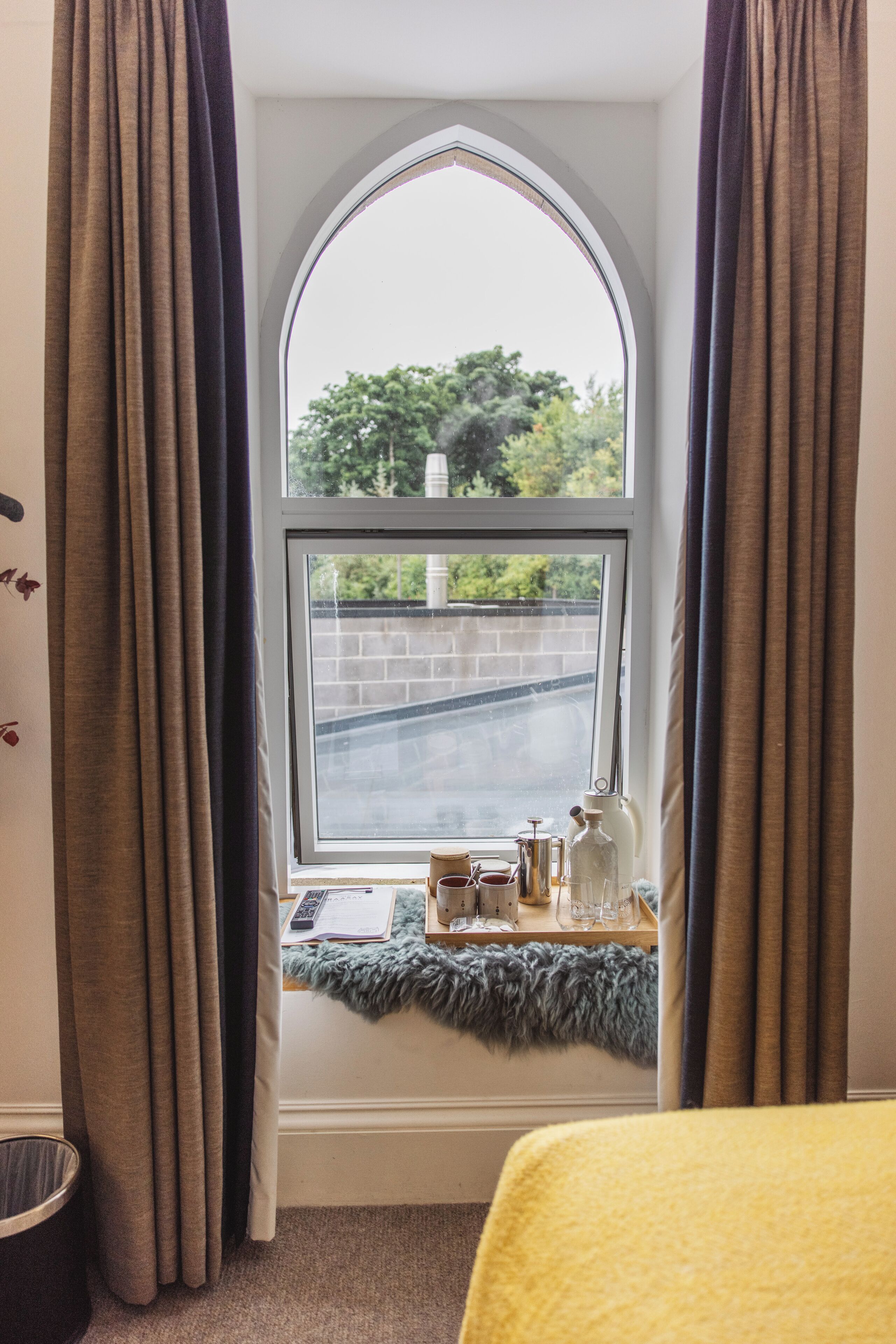 A room with a large arched window, curtains, a small table with a fur throw, and a view of a road and greenery outside.