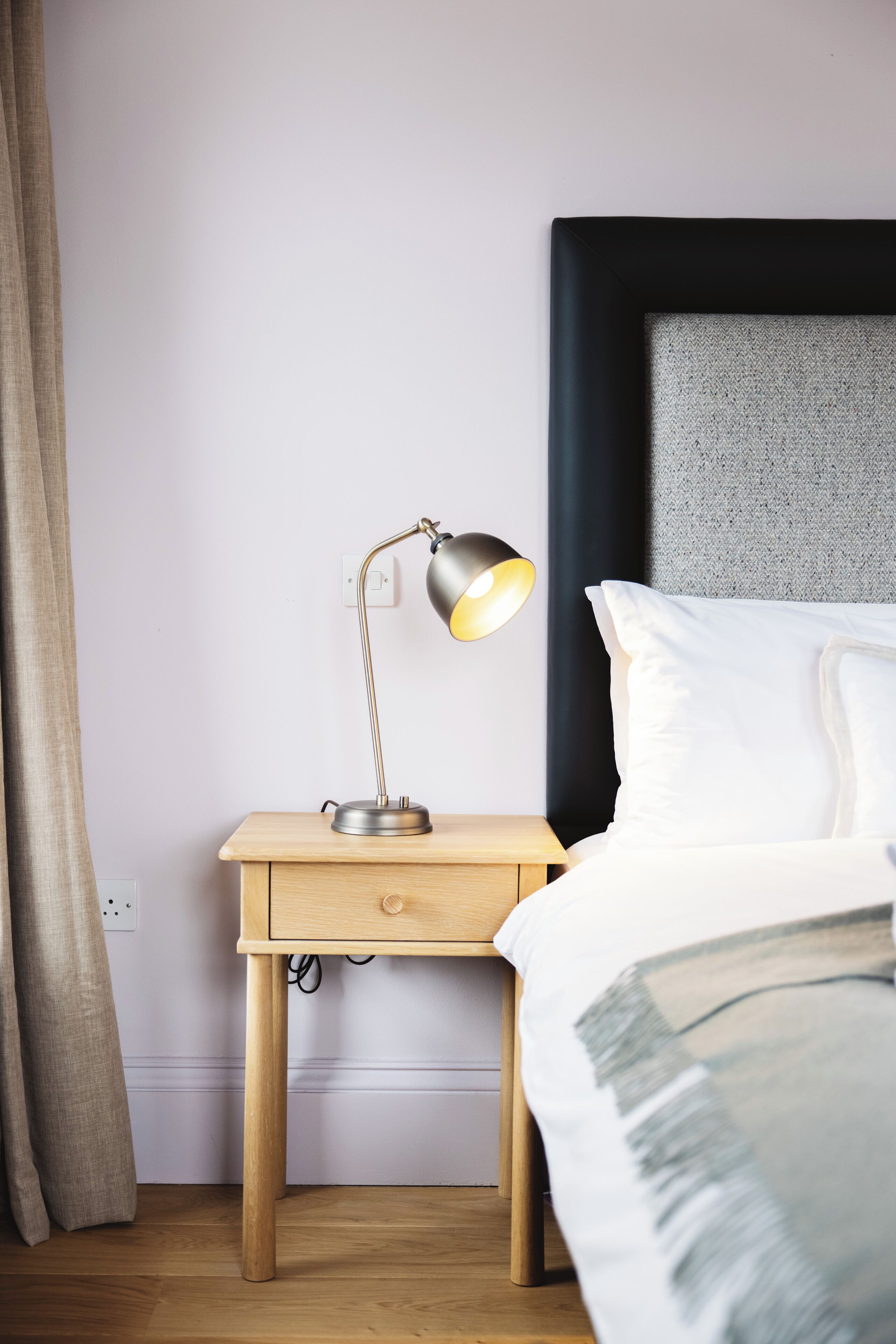 A neatly made bed with white linens, a bedside lamp on a wooden nightstand, and a plain wall with a power outlet.