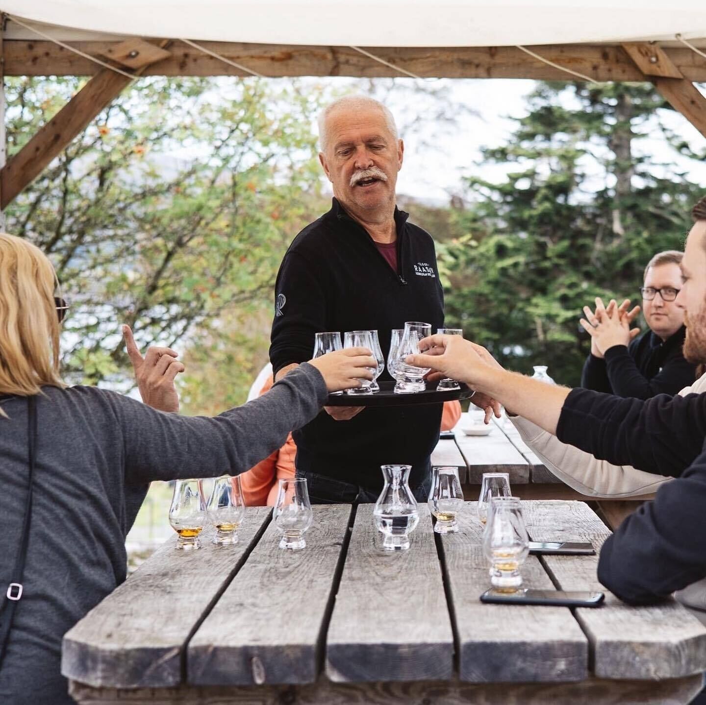 Un grupo de personas en una mesa de madera al aire libre, con vasos y botellas, bajo una estructura de madera.