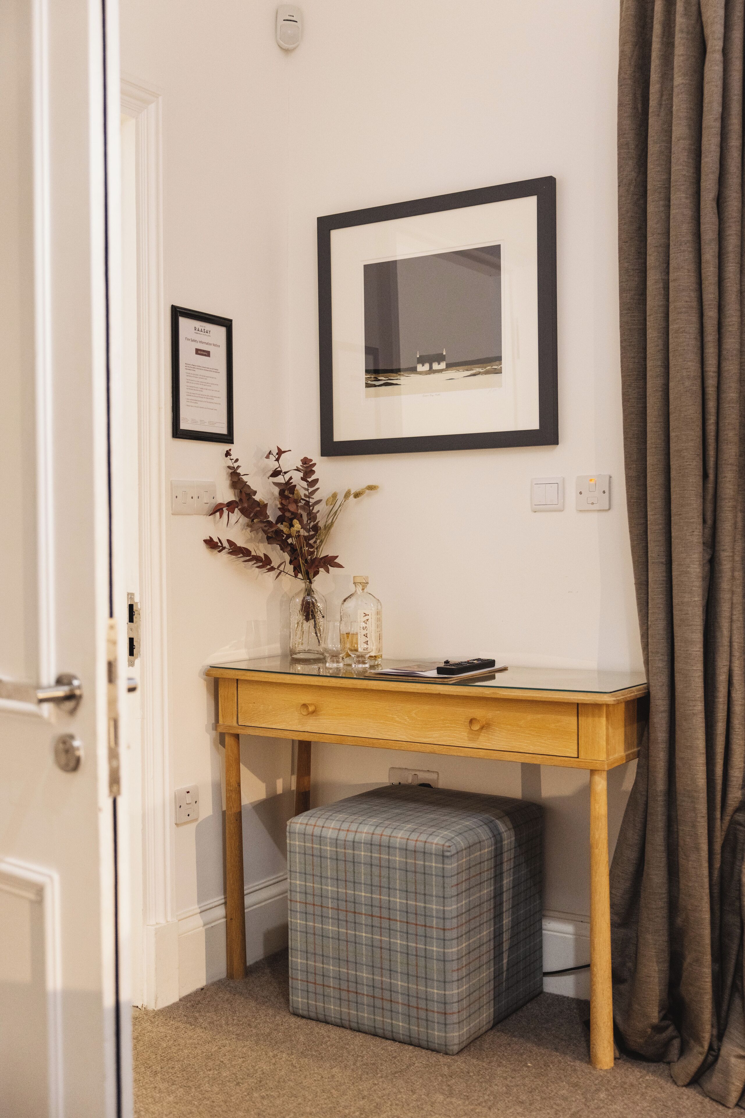 Une petite pièce, bien rangée, avec un bureau en bois, un miroir encadré, un vase contenant des fleurs séchées et un pouf à carreaux.