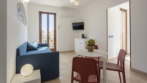 A modern living room with a blue sofa, a white table with red chairs, a flat-screen TV, and a city view. - Poseidon Residence (San Vito Lo Capo)