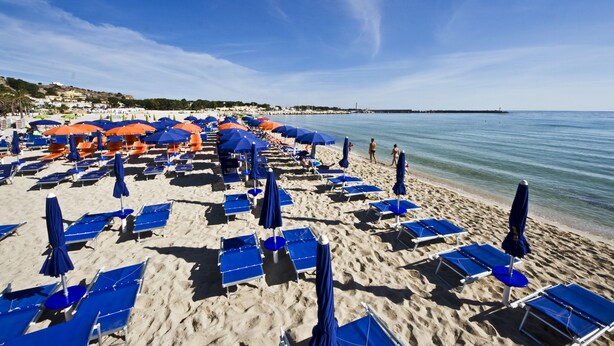 A beach with rows of blue and orange sun loungers and umbrellas, a sandy shore, and calm water. - Poseidon Residence (San Vito Lo Capo)