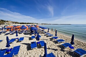 Una playa con filas de tumbonas y sombrillas azules y anaranjadas, una orilla arenosa y agua tranquila.