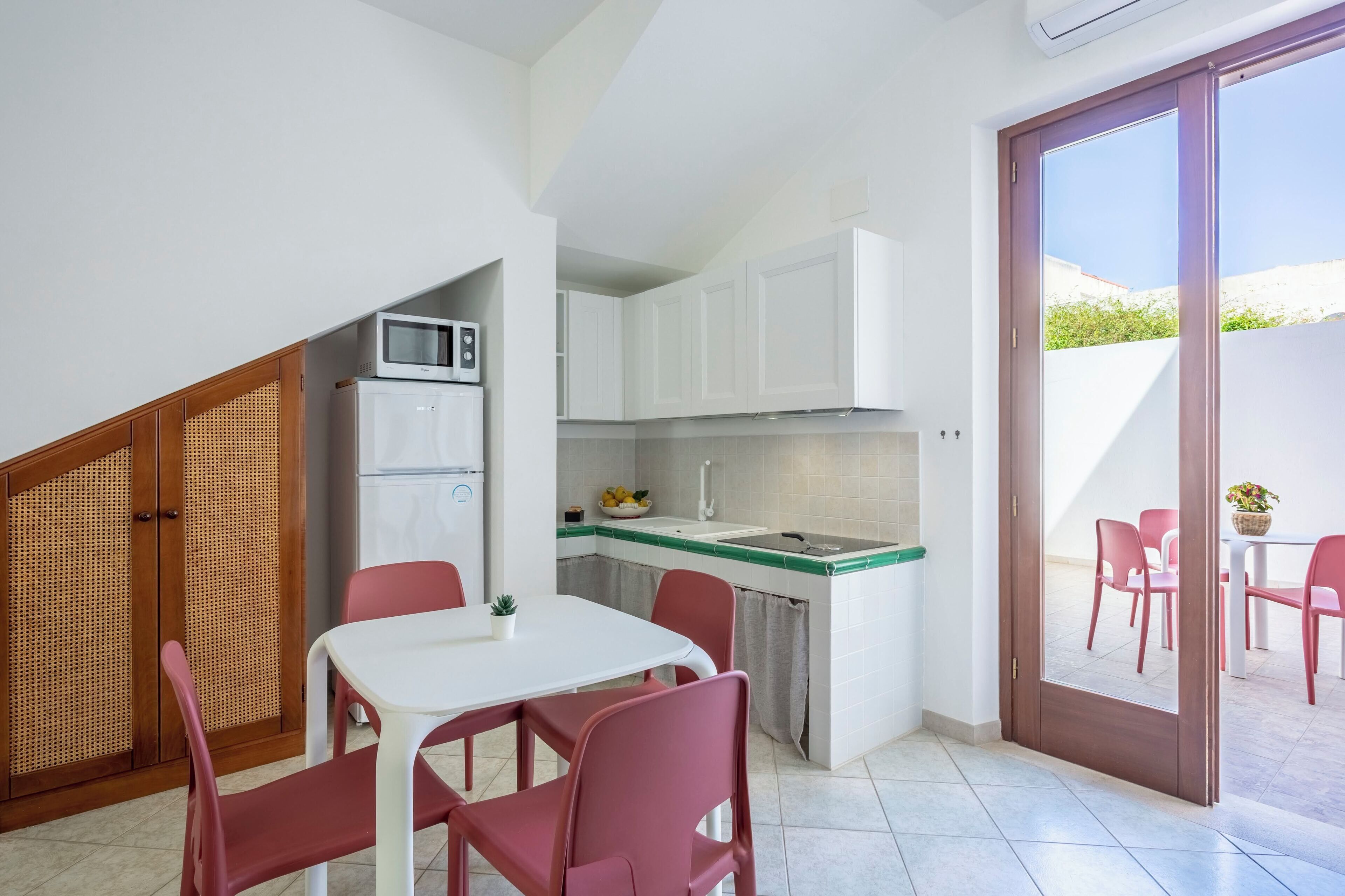 A compact kitchen with white cabinets, a microwave, and a sink. There is a dining area with a table and chairs, and a sliding glass door leading to an outdoor patio with chairs and a table.