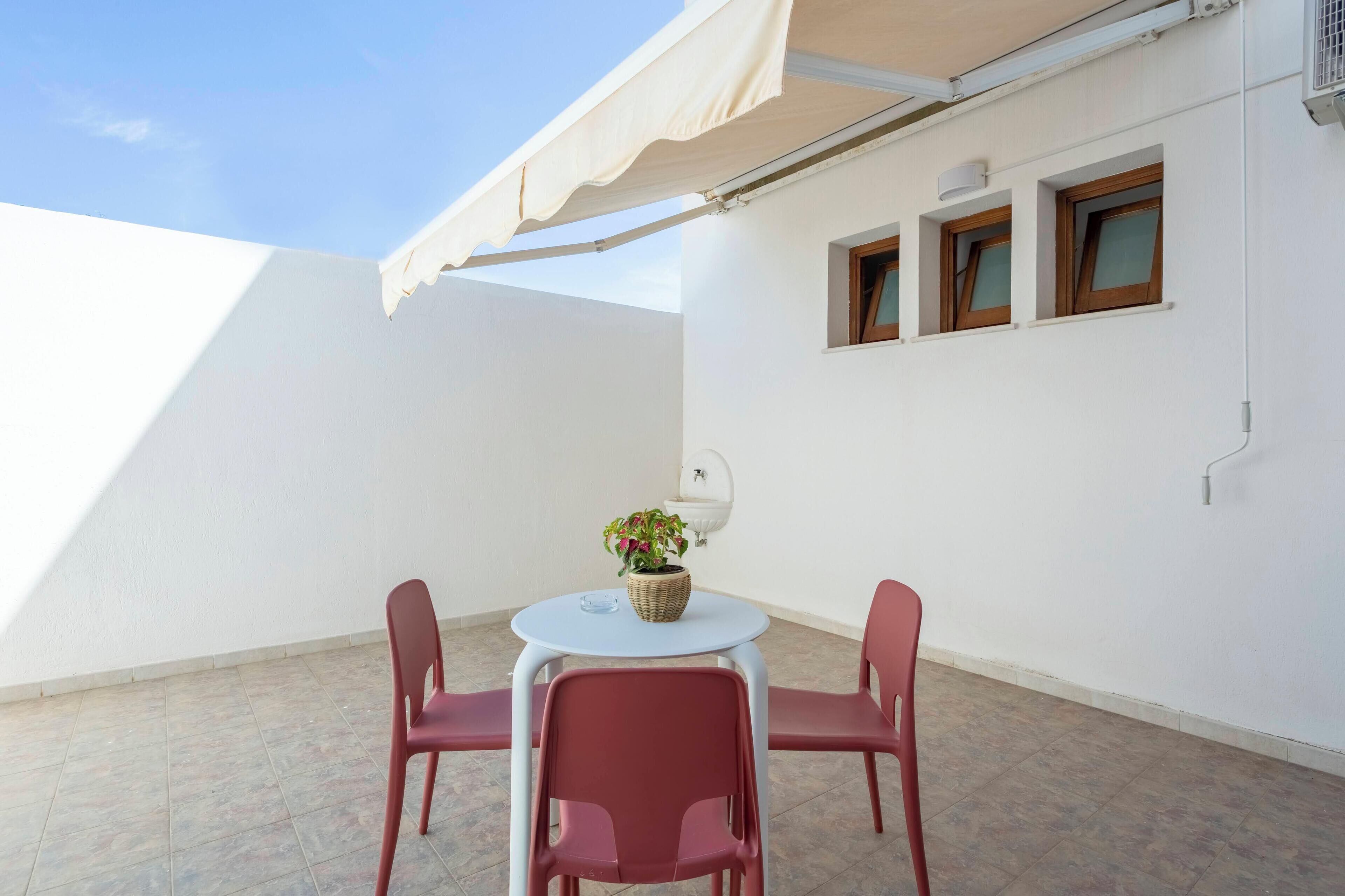 A small patio with a round table and two chairs, a potted plant, and a white wall with three windows.