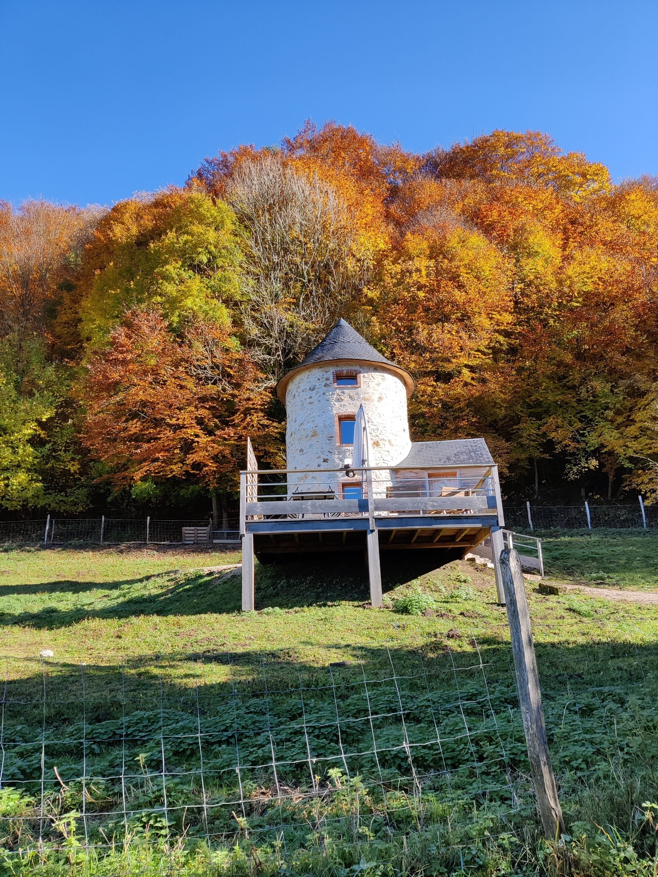 Una torre de piedra rústica con balcón, rodeada por un jardín cercado y follaje otoñal.