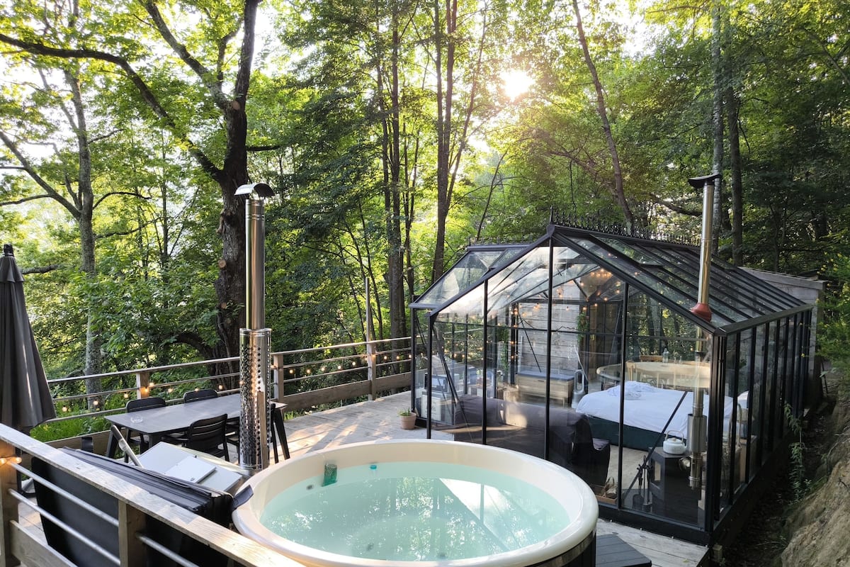 Un bain à remous doté d’un toit en verre et entouré d’une terrasse en bois, le tout niché au cœur d’une zone boisée, avec un espace de détente adjacent, également clos par des parois de verre et équipé de sièges.
