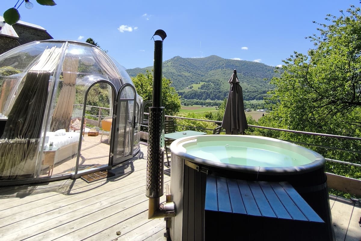 Une terrasse en bois avec un jacuzzi, un espace de couchage clos par des parois de verre, et une vue sur une vallée verdoyante et des montagnes.