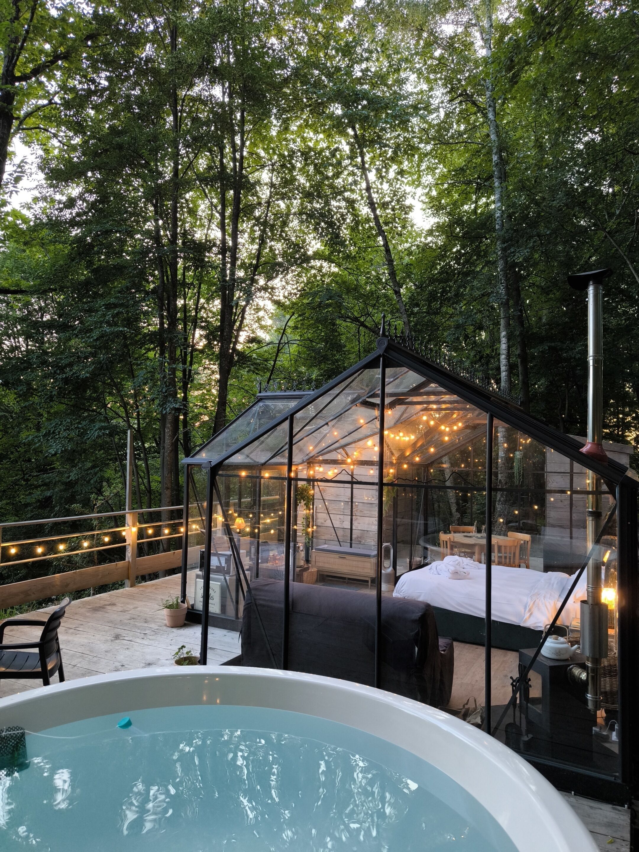 Eine Blockhütte mit Glasdach, einem Bett, einer Badewanne und Lichterketten in bewaldeter Landschaft.