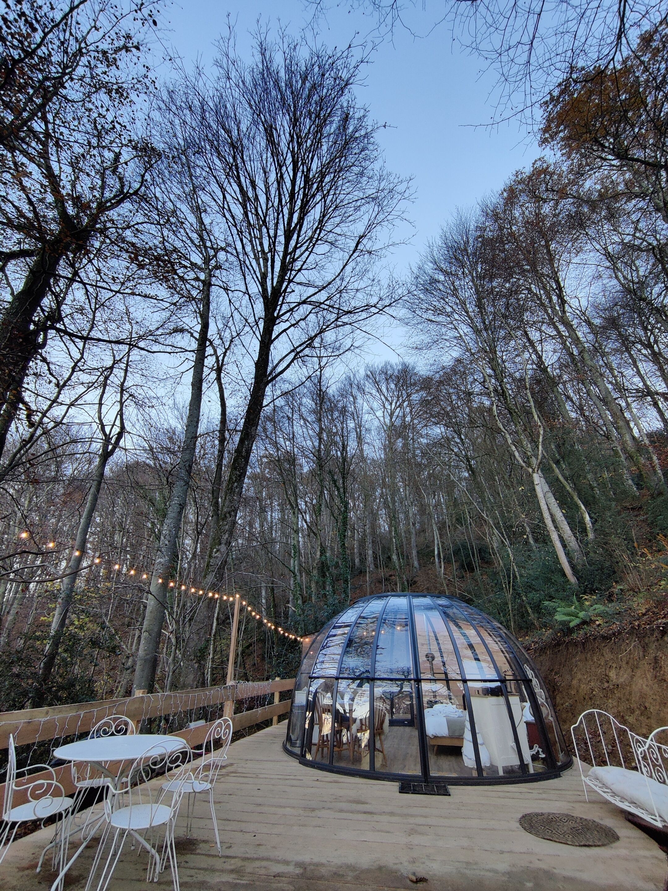 A transparent dome structure with a table and chairs inside, surrounded by a wooden deck and string lights.