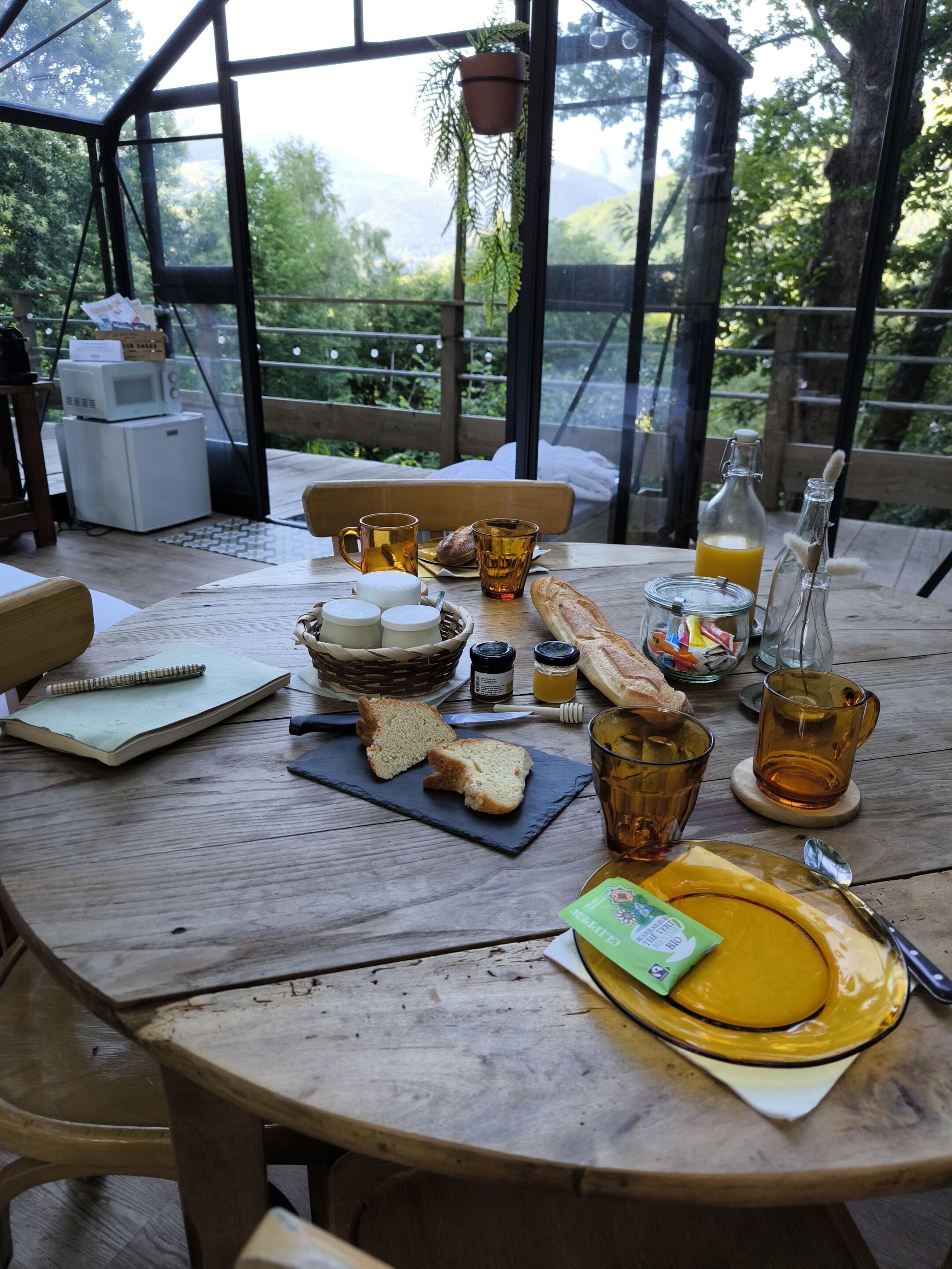 A wooden table set with breakfast items, including bread, butter, and honey, in a room with large windows and a view of greenery.
