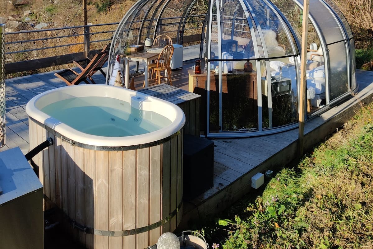 Un jacuzzi entouré de parois en verre, avec une terrasse en bois et un espace salon.