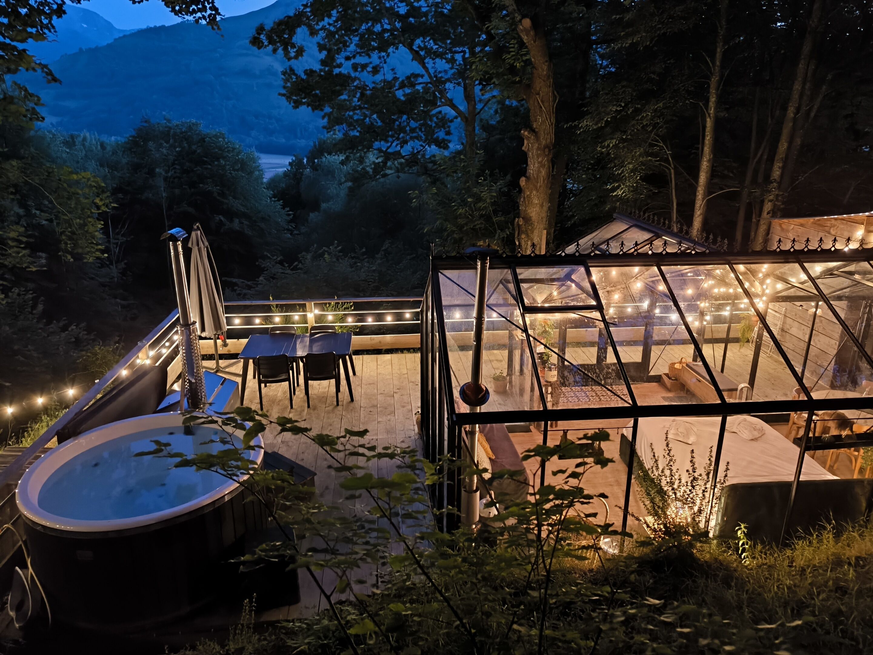 A cozy outdoor patio with a hot tub, a dining table, and a glass-enclosed area with string lights.