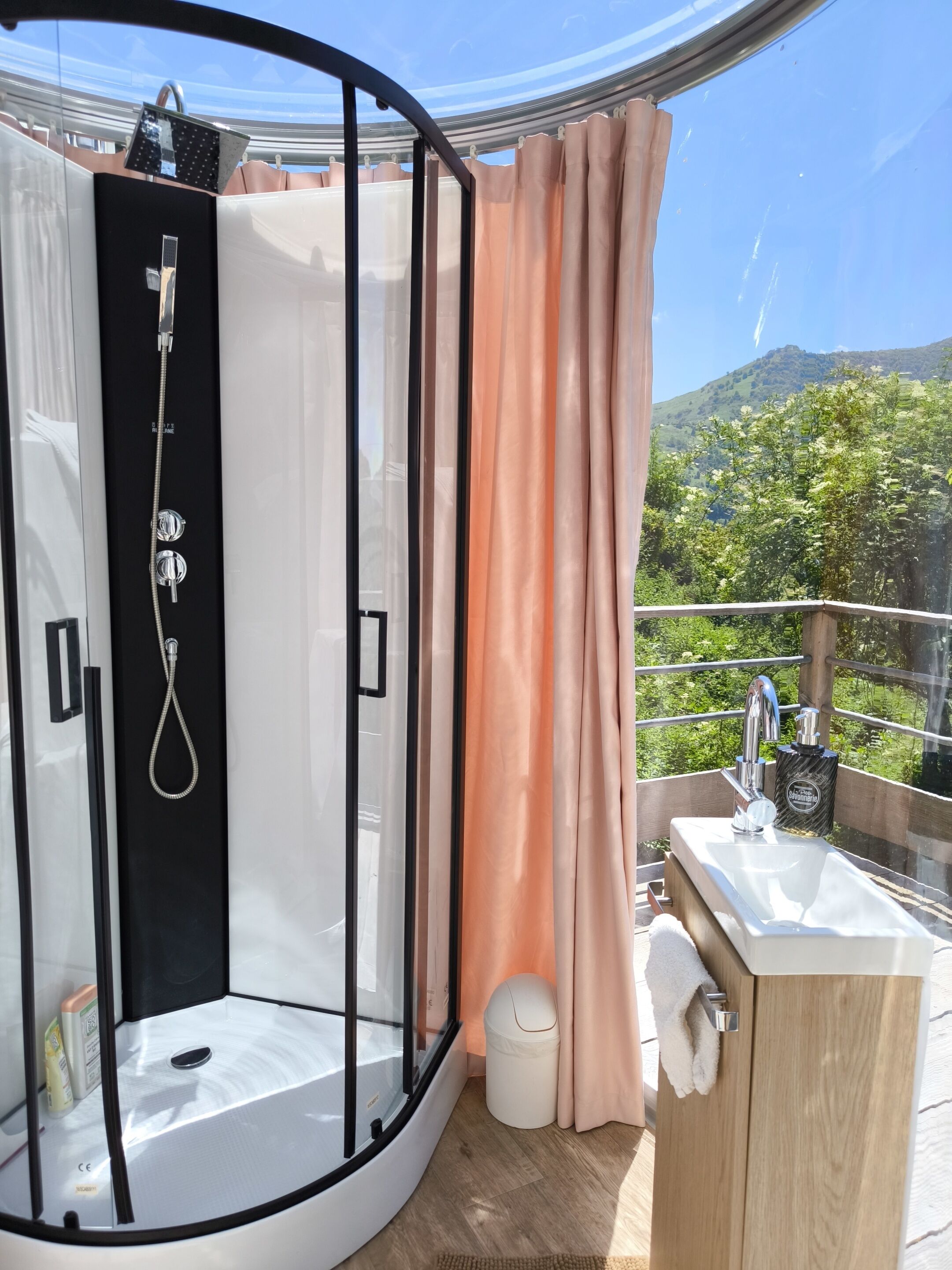Ducha con cerramiento de vidrio, cabezal de ducha de mano, lavamanos con grifo y un pequeño cubo de basura.