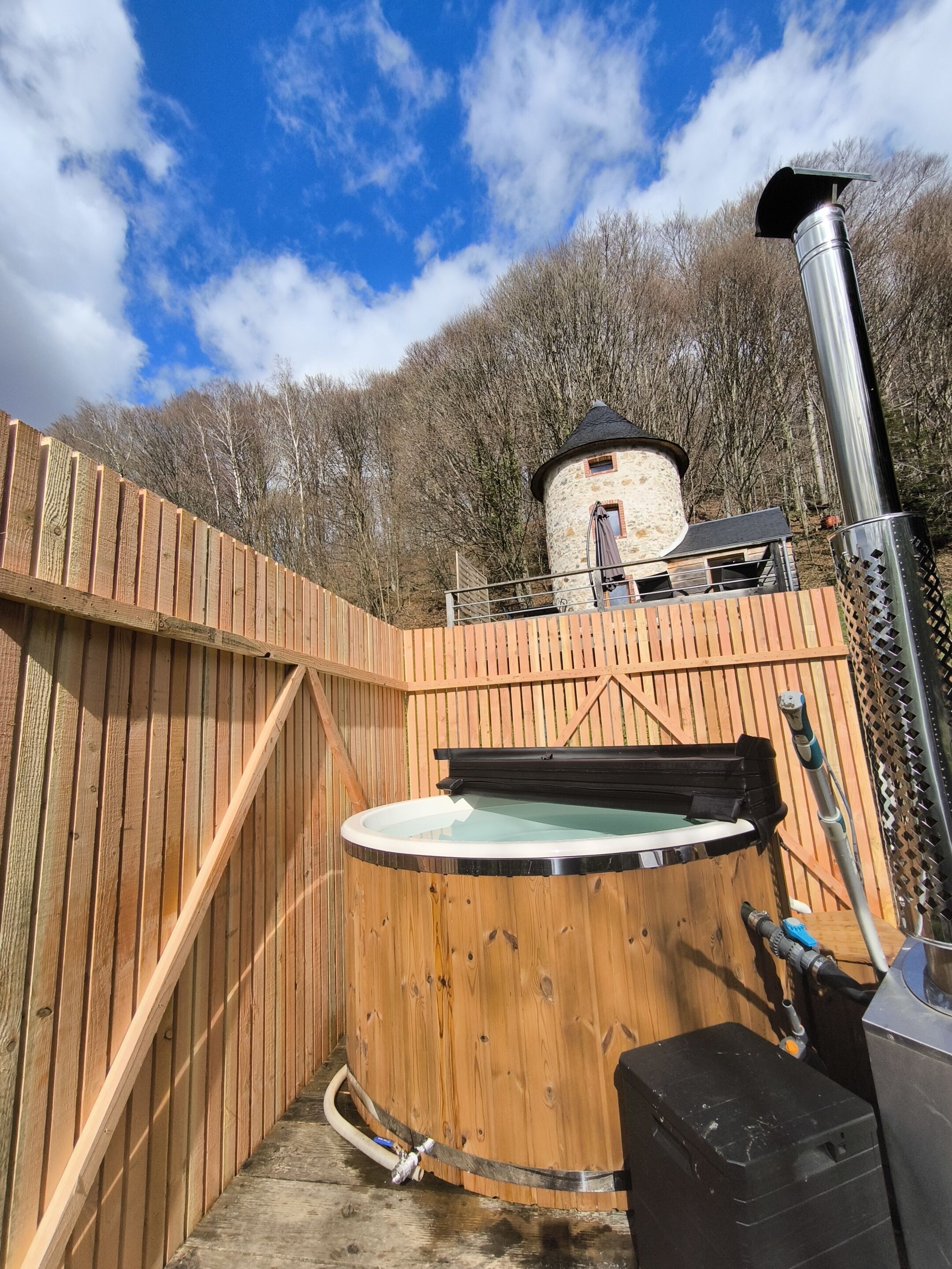 A wooden hot tub with a stone structure in the background, surrounded by a wooden fence.