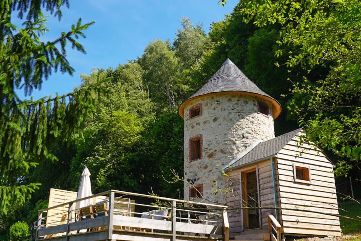 Une tour rustique en pierre, surmontée d’un toit conique, dotée d’une terrasse en bois et d’une petite cabane attenante.