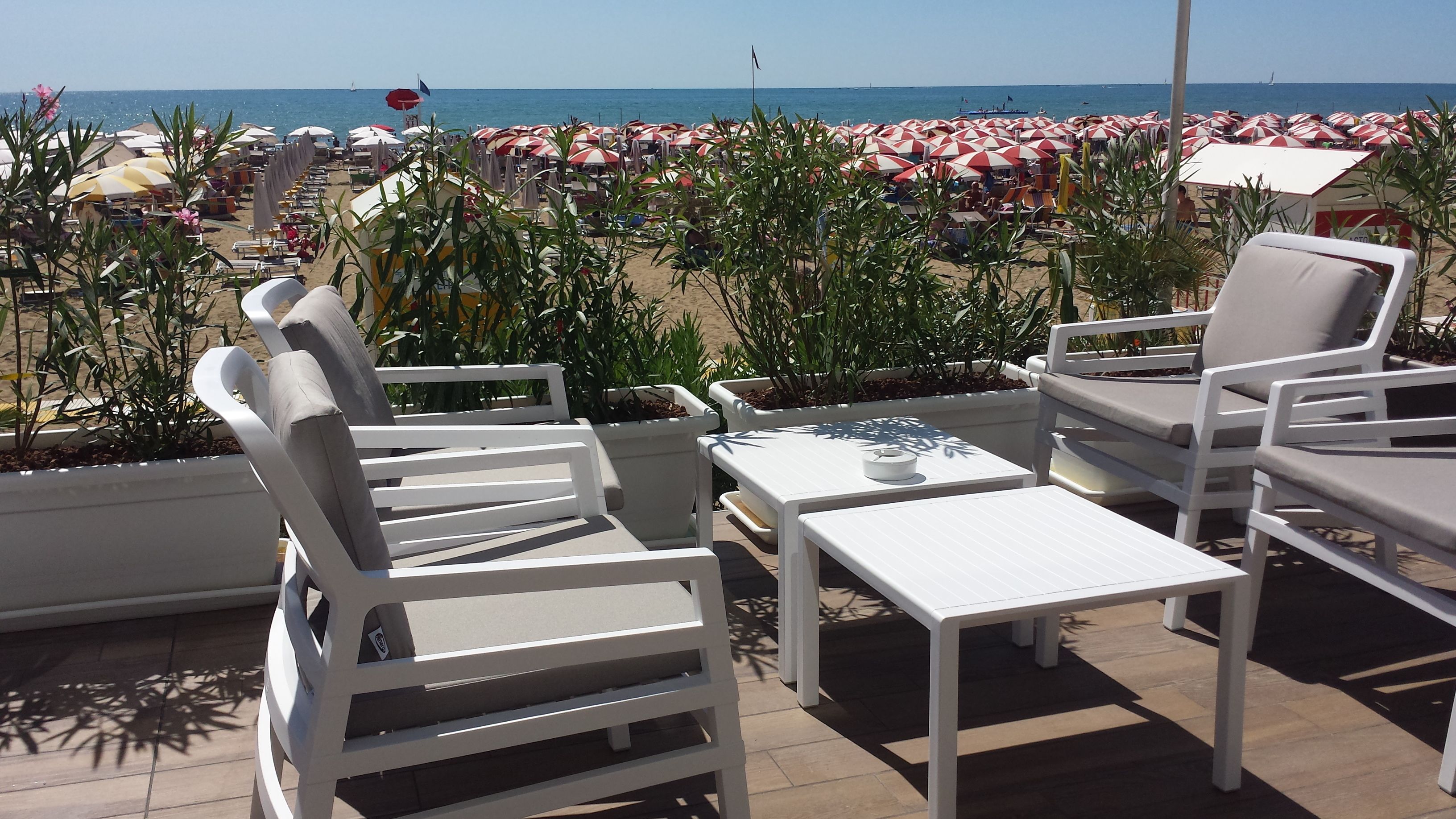 A terrace with white lounge chairs and a table, overlooking a beach with red and white umbrellas.