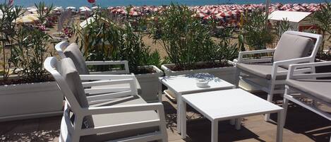 A terrace with white lounge chairs and a table, overlooking a beach with red and white umbrellas.