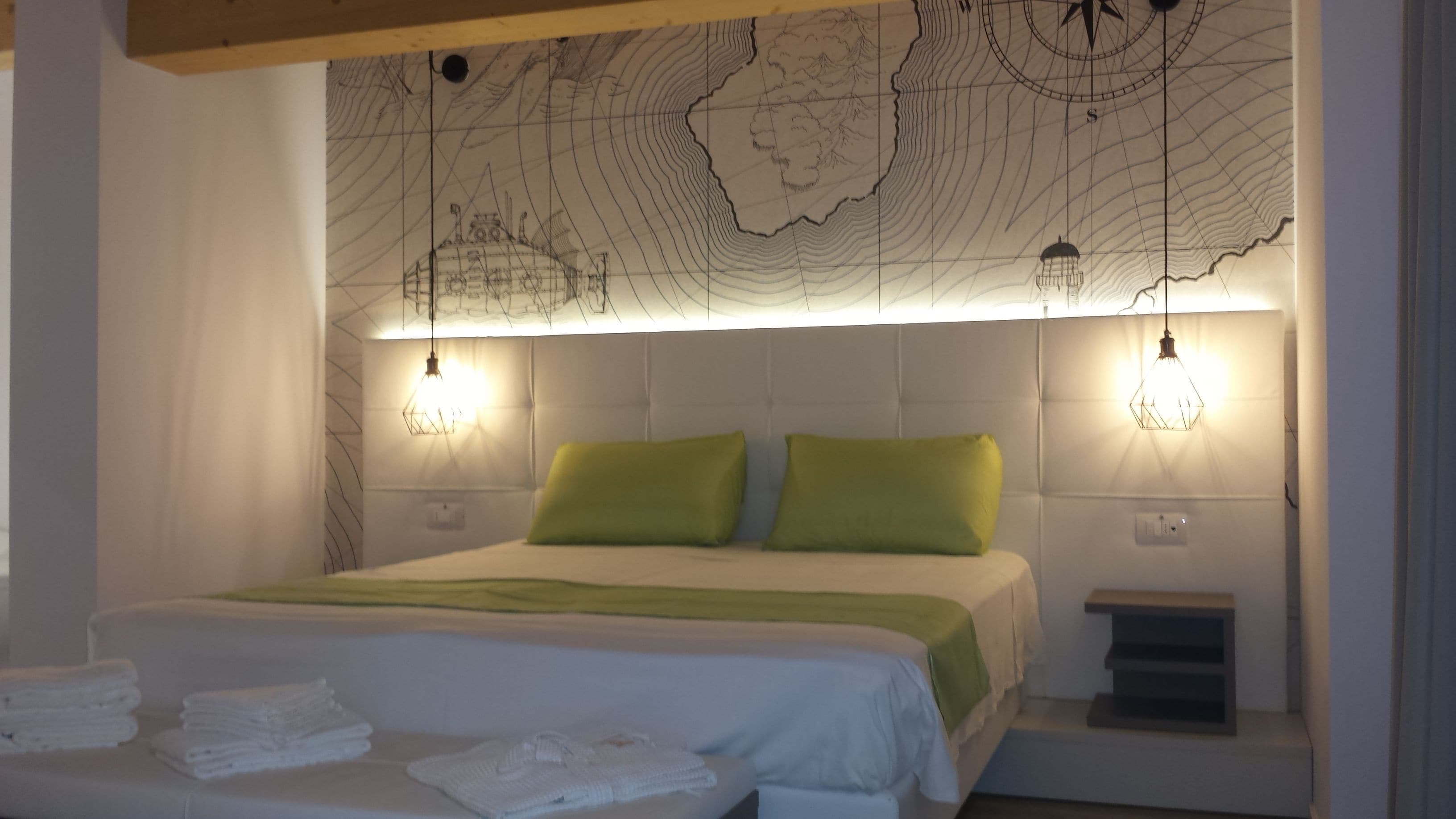 A bedroom with a bed, green pillows, a map wall, two wall-mounted lights, and a nightstand.