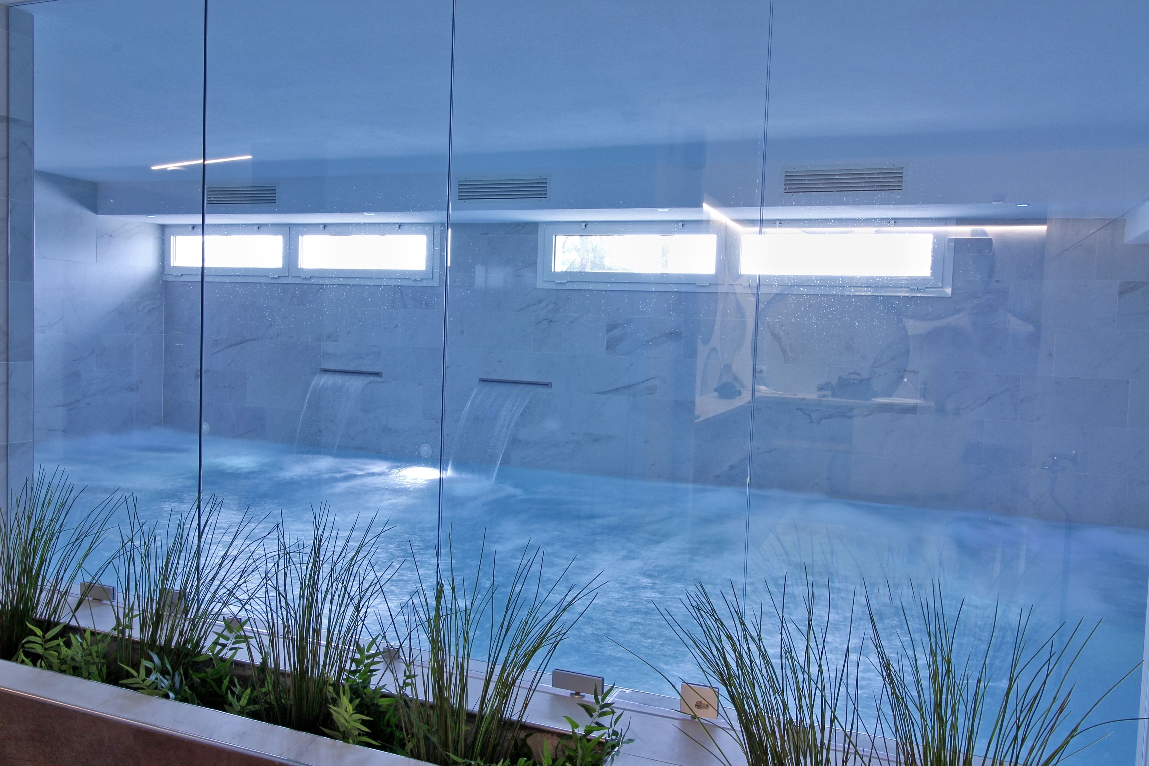 Une piscine intérieure, entourée de plantes, avec des parois en verre offrant une vue dégagée sur le ciel et dont l’eau est cristalline.