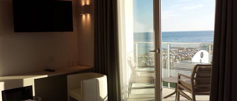 Habitación de hotel con cama, escritorio, silla, televisor montado en la pared y balcón con vista a la playa y al mar.