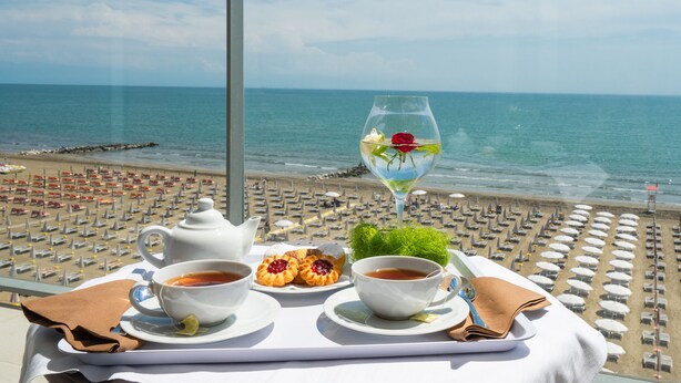 A table with tea, pastries, and a glass of water on a balcony overlooking a beach with rows of umbrellas. - Hotel Elite (Caorle)
