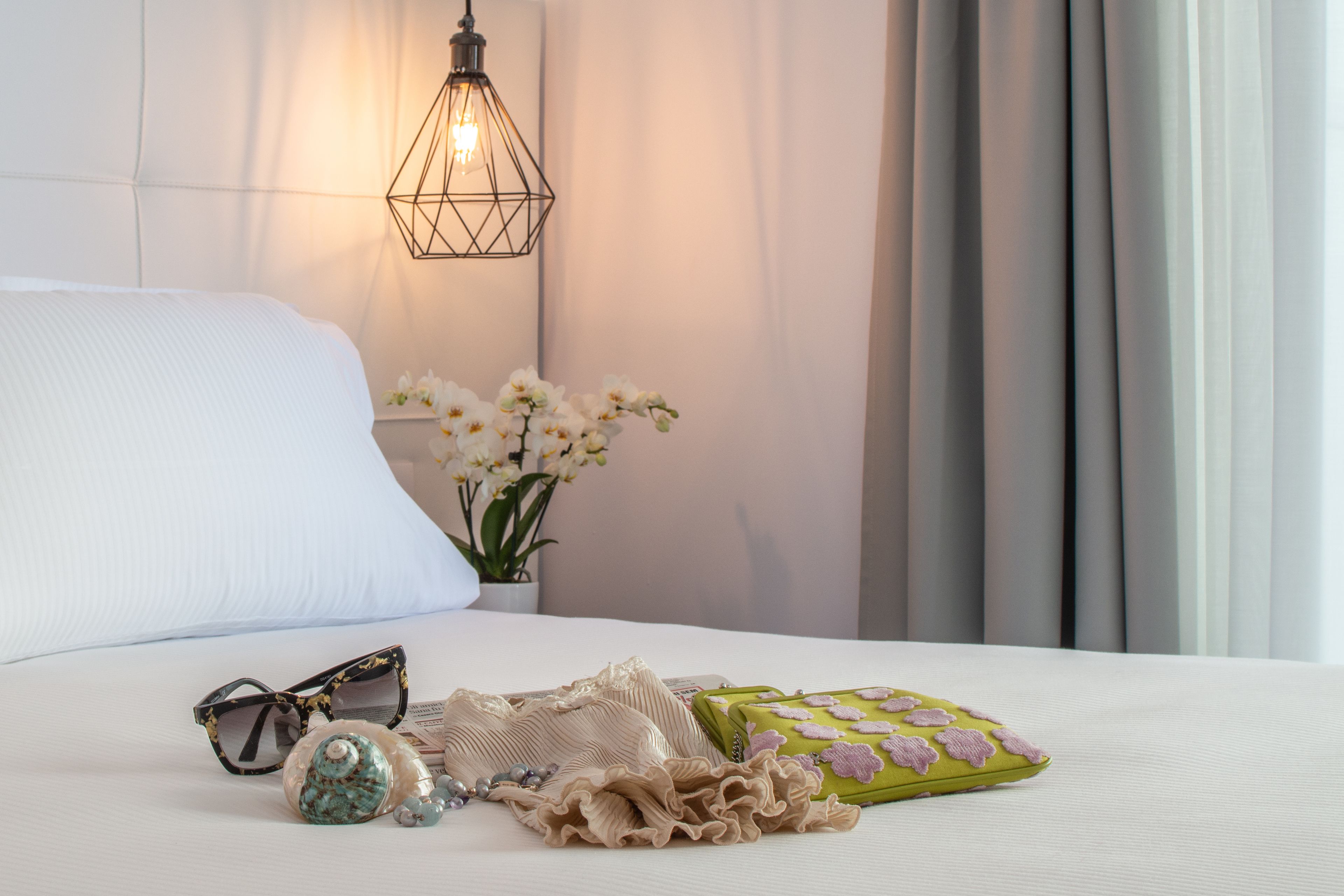 Una cama bien tendida con una almohada blanca, una lámpara decorativa, un jarrón con flores blancas y una almohada verde con motivos florales.