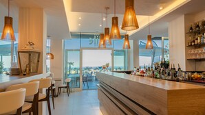A modern bar area with a wooden counter, multiple hanging pendant lights, and a view of the sea through large windows. - Hotel Elite (Caorle)