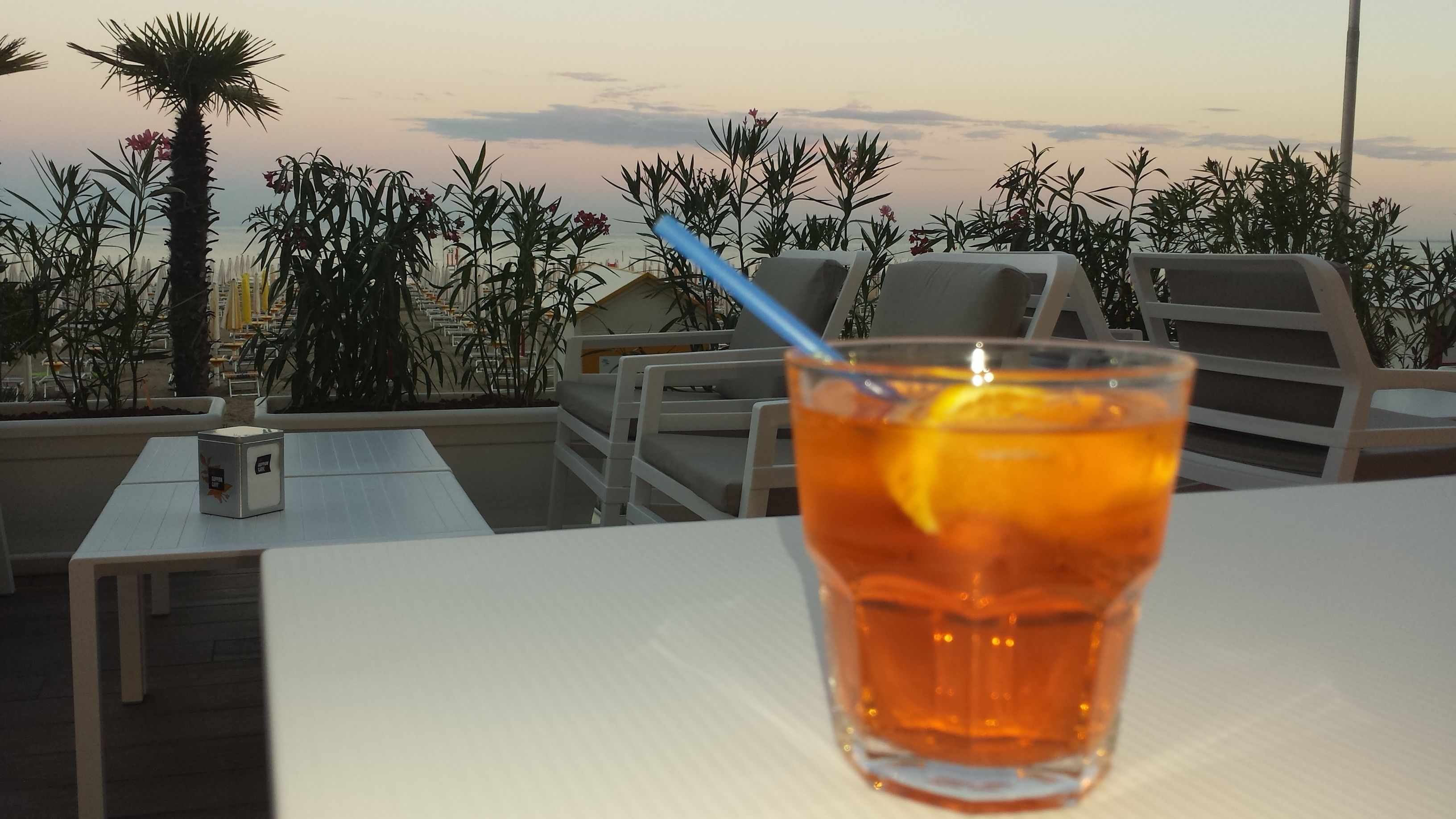 Un vaso de bebida color ámbar con una rodaja de limón y una pajita sobre una mesa blanca, con vista a una playa con tumbonas y palmeras.