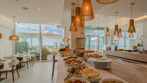 A modern dining area with a buffet setup, large windows offering a view of the sea, and contemporary pendant lighting. - Hotel Elite (Caorle)