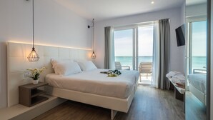 A modern hotel room with a large bed, a small bedside table with a plant, a TV mounted on the wall, and a balcony with a view of the sea. - Hotel Elite (Caorle)