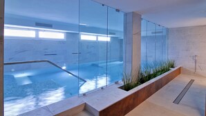 A modern indoor swimming pool with glass walls, a stone-tiled floor, and a wooden planter with greenery. - Hotel Elite (Caorle)