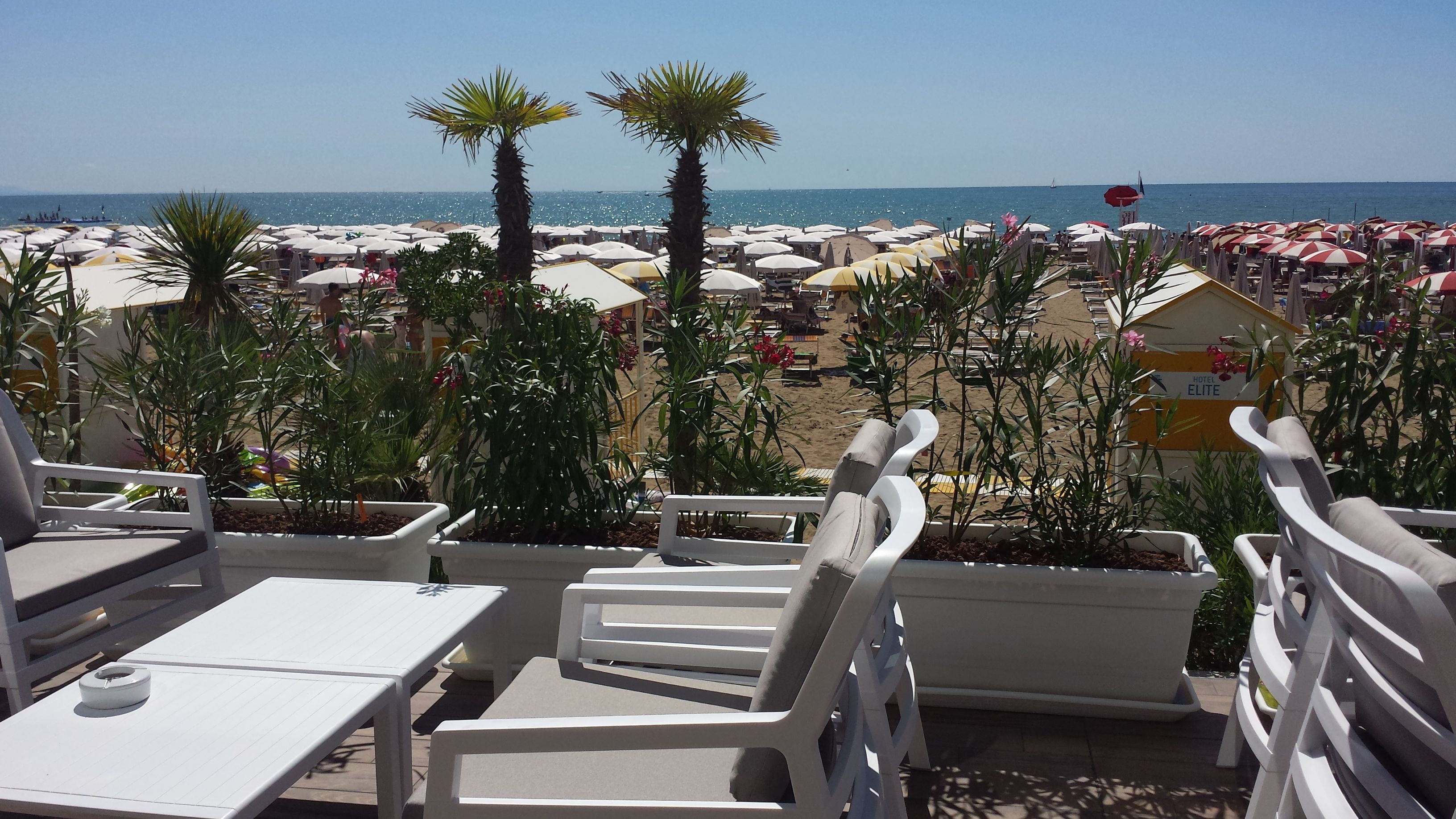 Zona relax in spiaggia con poltrone bianche e tavolini, vista su una spiaggia con ombrelloni e palme.