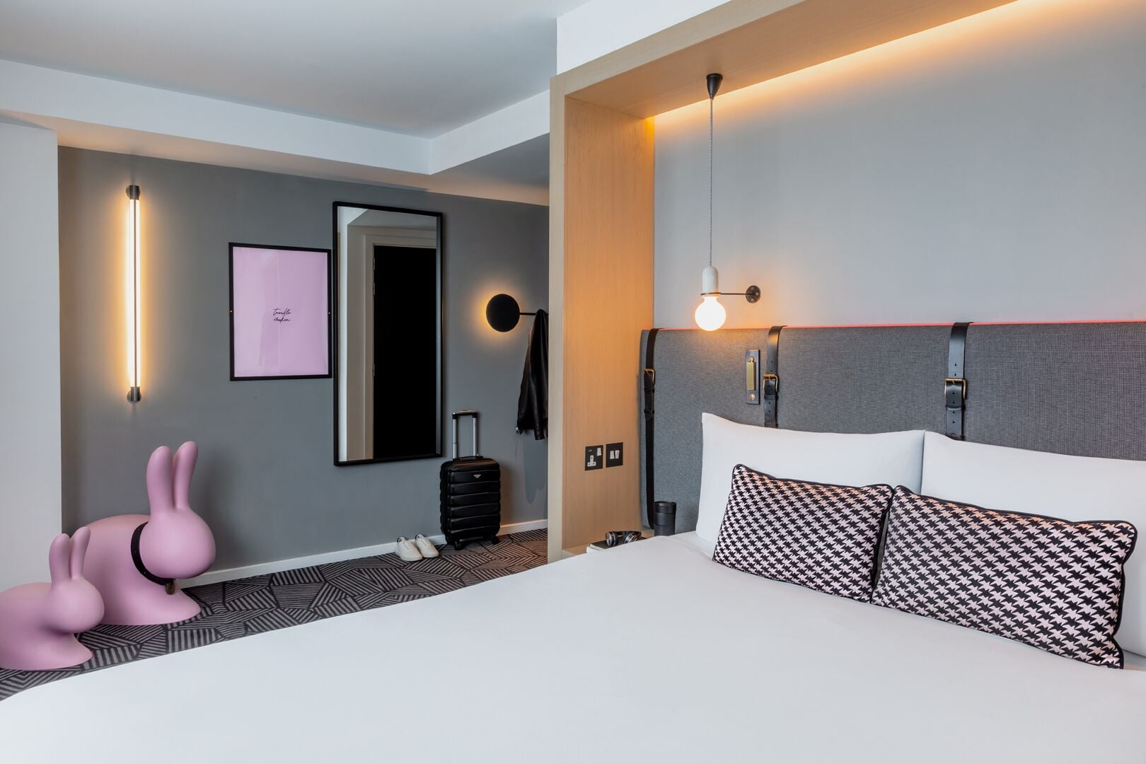A modern hotel room with a large bed, a suitcase, a pink bunny decoration, and a mirror.