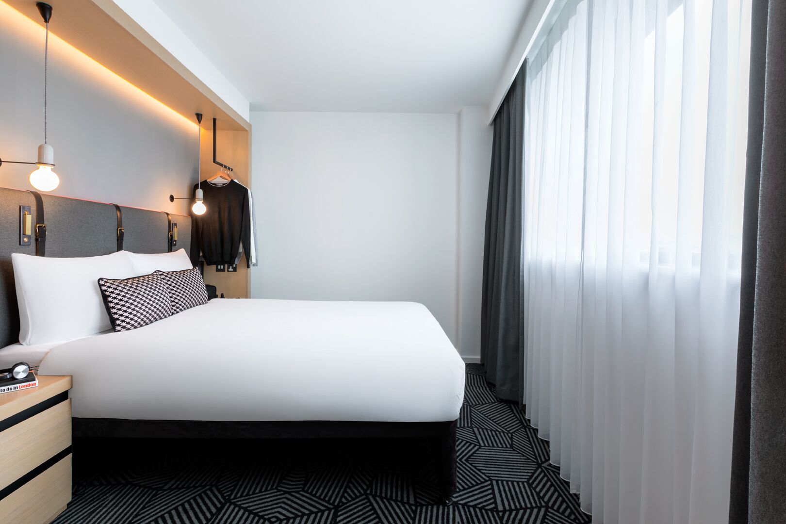 Ein modernes Hotelzimmer mit einem großen Bett, einem Nachttisch, einem Kleiderhaken und einem Fenster mit Vorhängen.