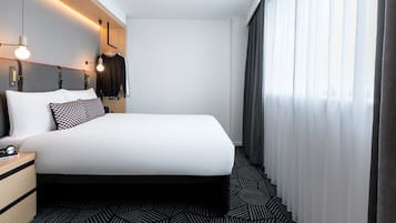 A modern hotel room with a large bed, a nightstand, a hanging coat rack, and a window with curtains.