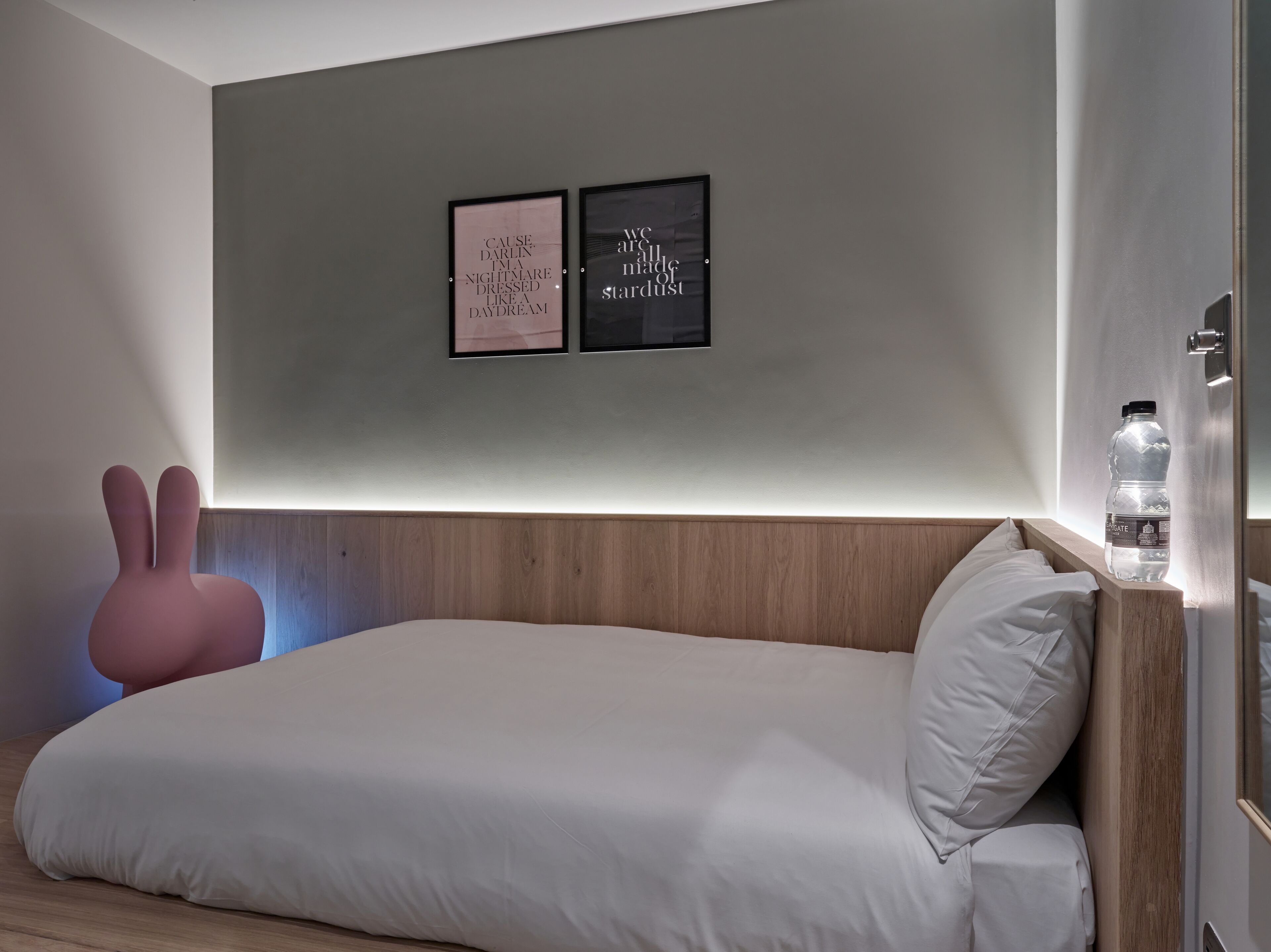 Un dormitorio moderno con cabecera de madera, cama blanca, almohada con forma de conejo rosa y dos citas enmarcadas en la pared.