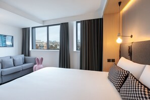 A modern hotel room with a large bed, a grey sofa, a pink chair, and a city view through the windows. - Assembly Leicester Square (London)