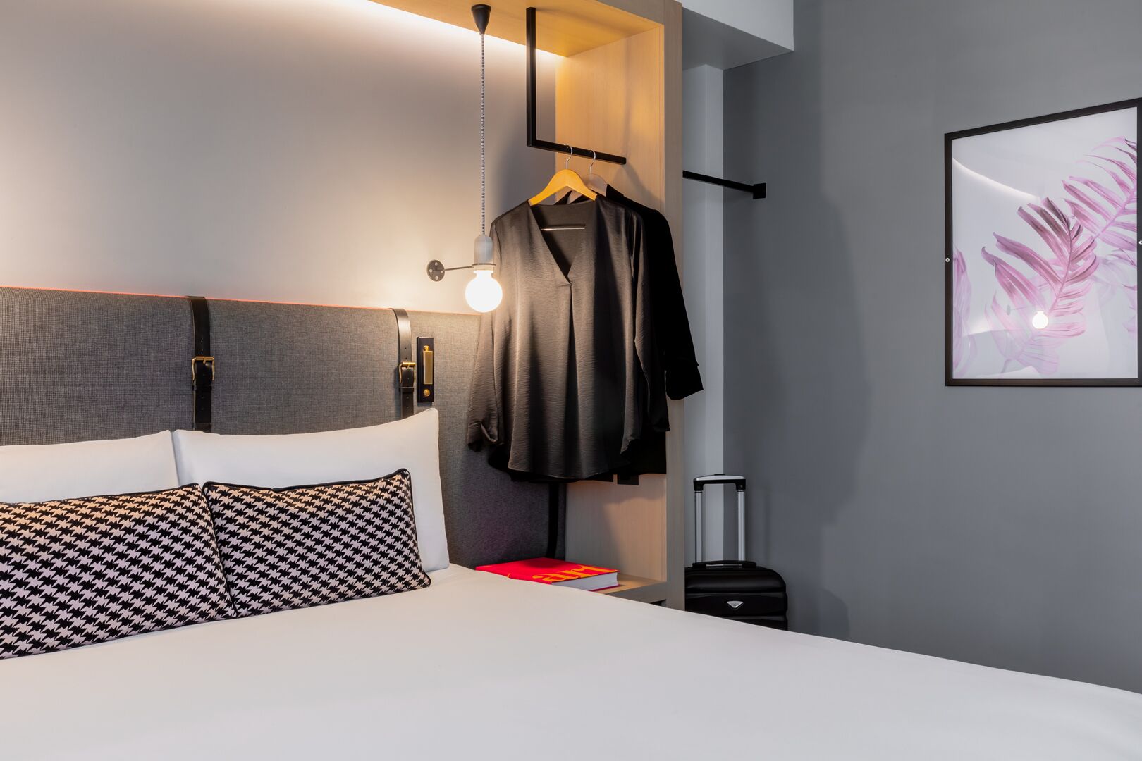 Een moderne hotelkamer met een bed, een kapstok, een koffer en een lijst met kunstwerk aan de muur.