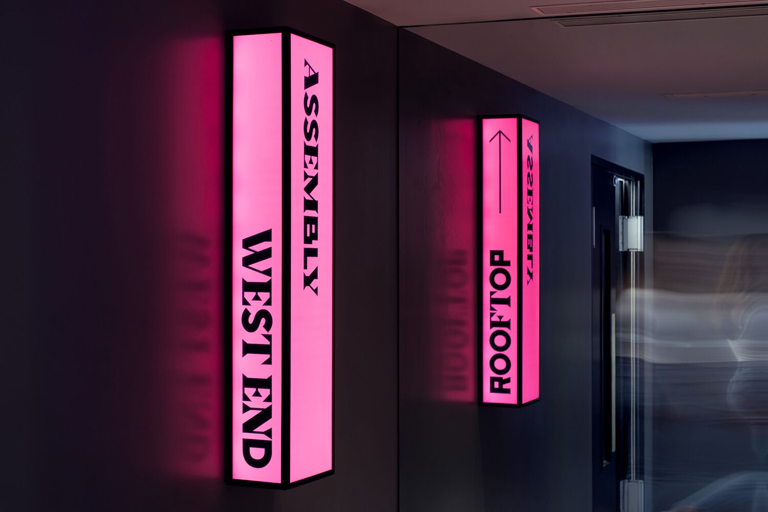 Eine Wand mit beleuchteten Schildern, die Raumbezeichnungen zeigen: 'ASSEMBLY', 'WESTIND' und 'ROOFTOP'.