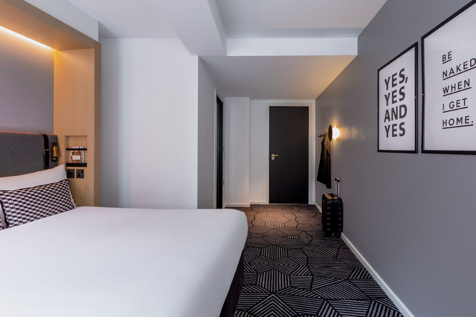 A hotel room with a large bed, a geometric patterned carpet, a black door, and motivational wall art.