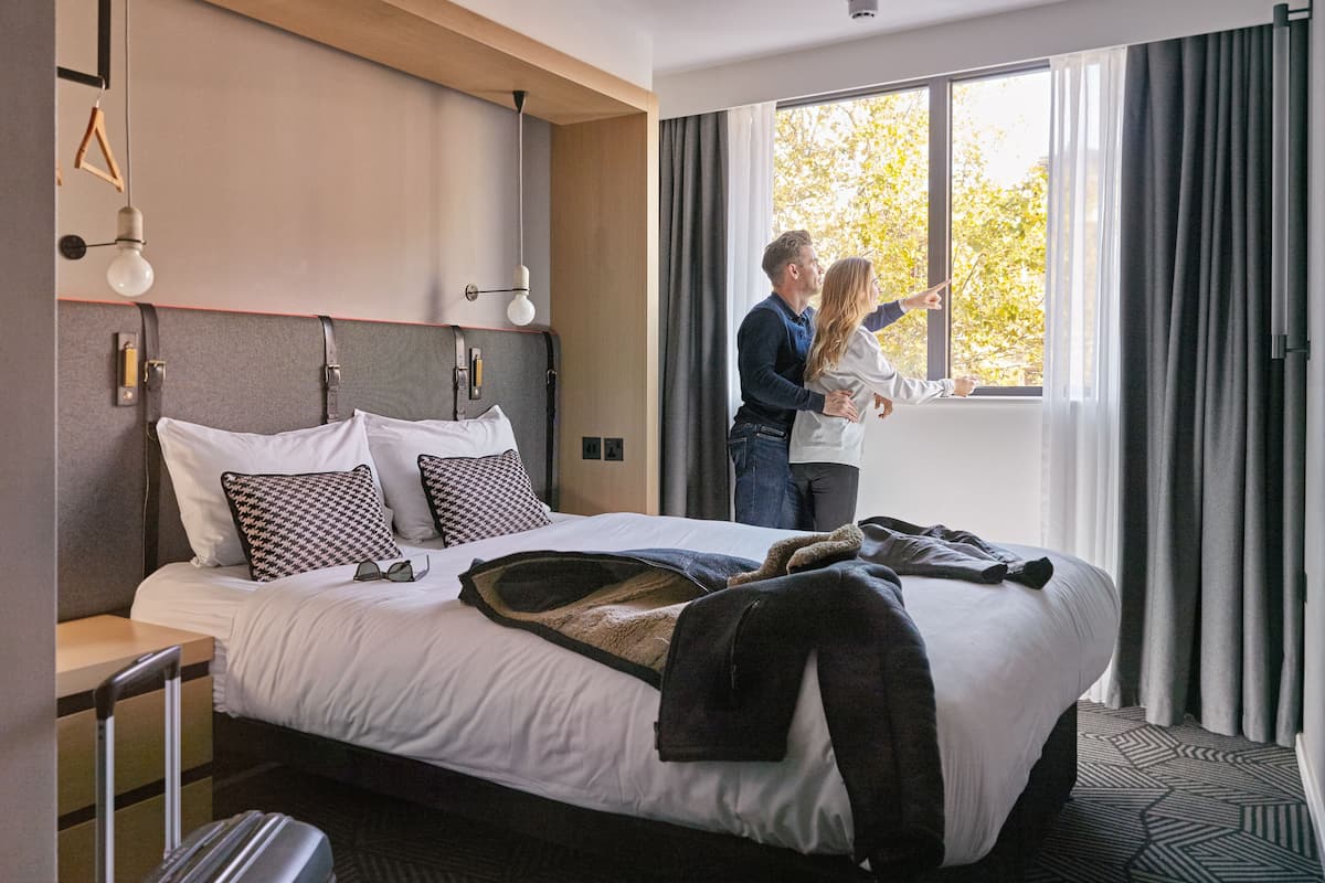 A modern hotel room with a large bed, a bedside table, a lamp, and a view of trees through the window.