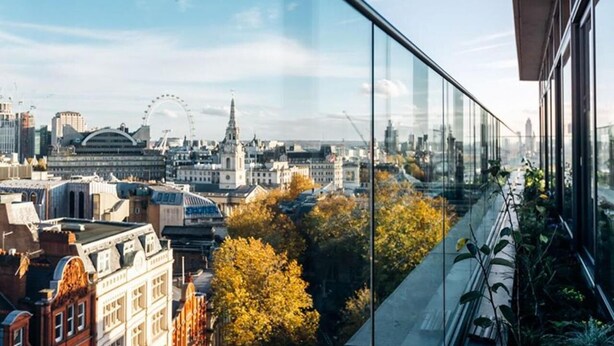 A cityscape view from a high-rise building with a glass railing, featuring historic architecture, a prominent tower, and a large Ferris wheel in the distance.