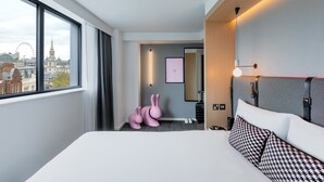 A hotel room with a large bed, a view of the city, a mirror, a wall-mounted light, and a pink bunny sculpture.
