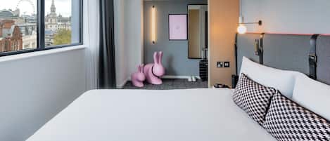 A hotel room with a large bed, a view of the city, a mirror, a wall-mounted light, and a pink bunny sculpture.