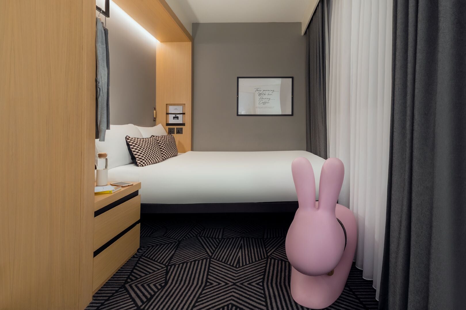 A hotel room with a bed, a wooden wardrobe, a bedside table, a pink chair, and a framed quote on the wall.