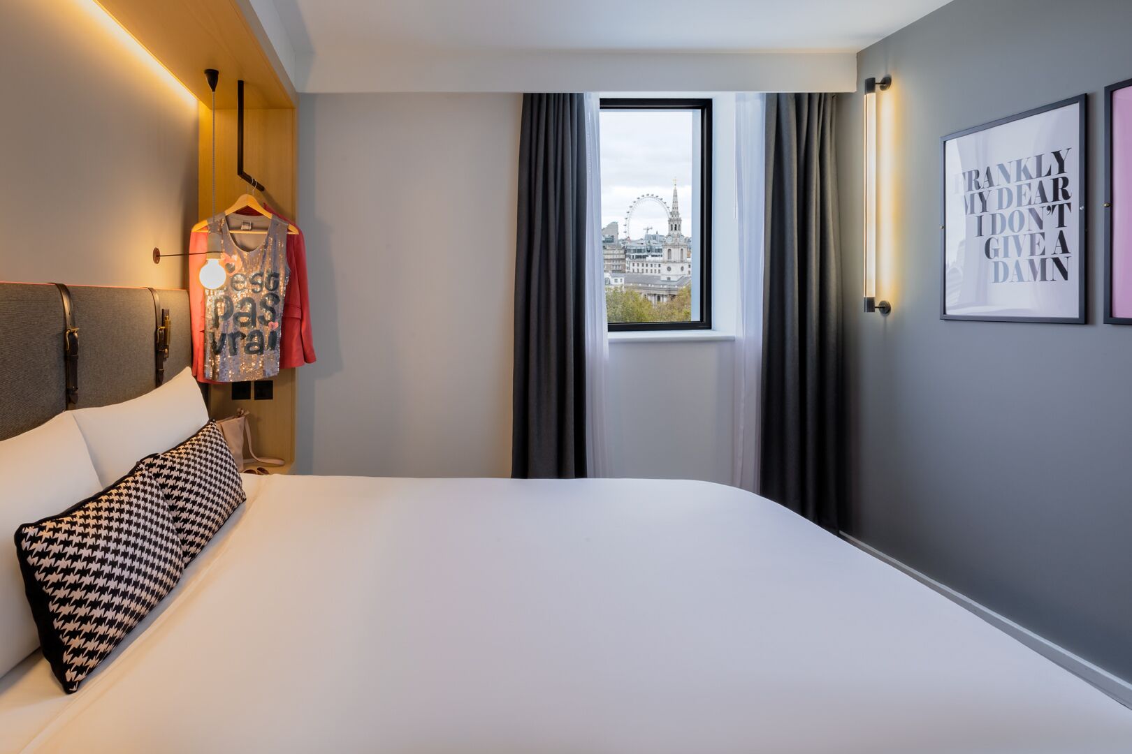 A hotel room with a large bed, a view of a cityscape through a window, and a wall-mounted light fixture.