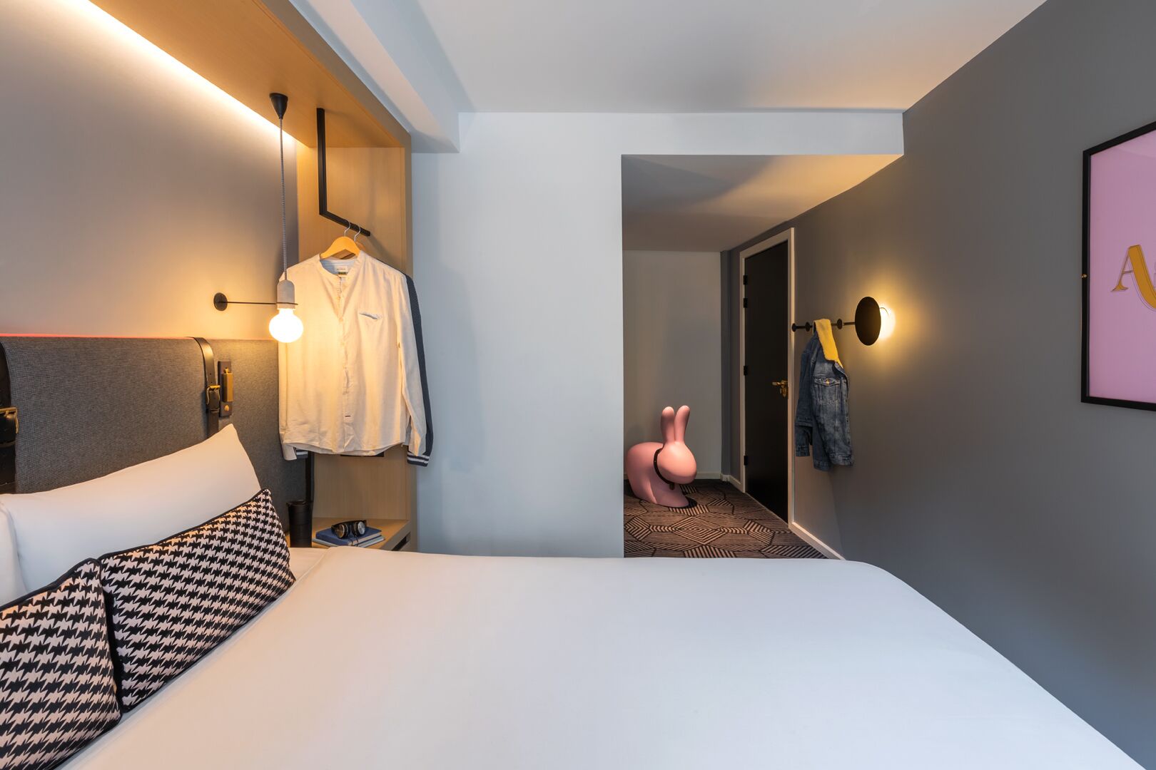 Een moderne hotelkamer met een groot bed, een opbergmantel, een opvallende roze konijnenstoel en een ingelijst schilderij aan de muur.