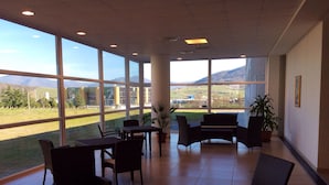 A spacious indoor area with large windows offering a view of a green landscape and mountains. There are several wicker chairs and a table, with potted plants adding greenery. - Hotel Residence Santa Loja (Tito)